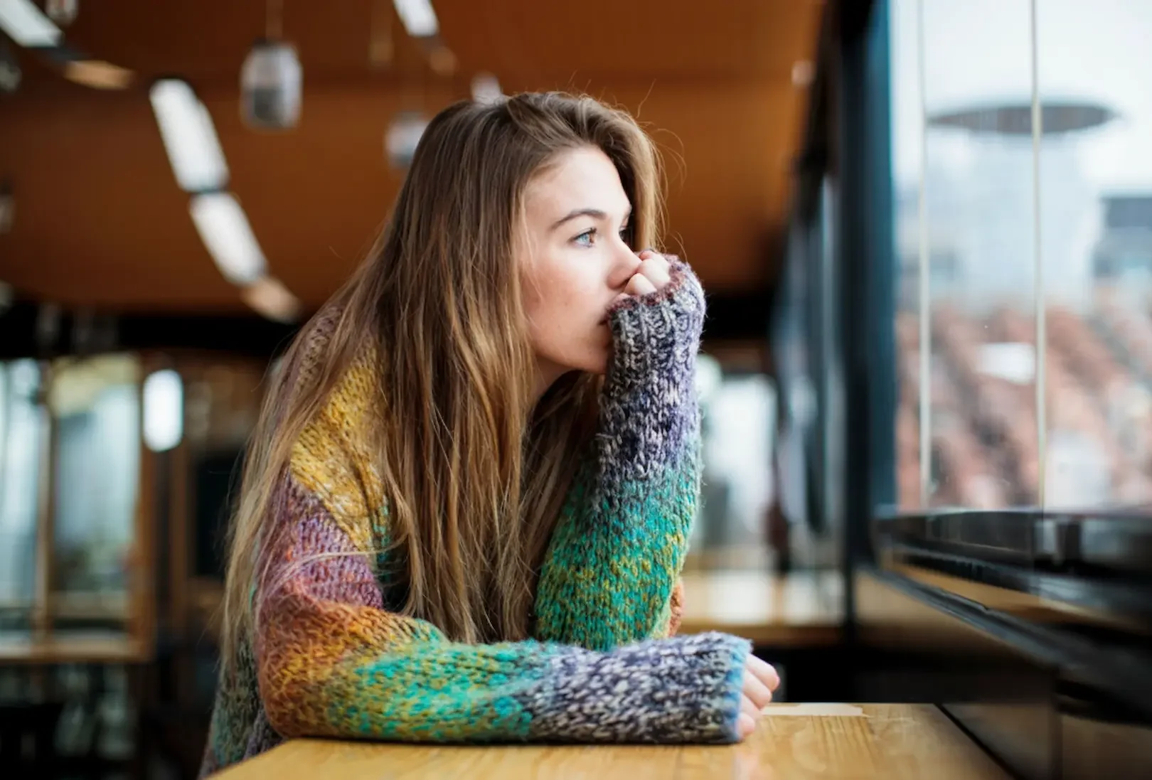 A young woman in a colorful sweater looking thoughtfully out a cafe window, depicting the emotional isolation often found in the depression vs anxiety debate.