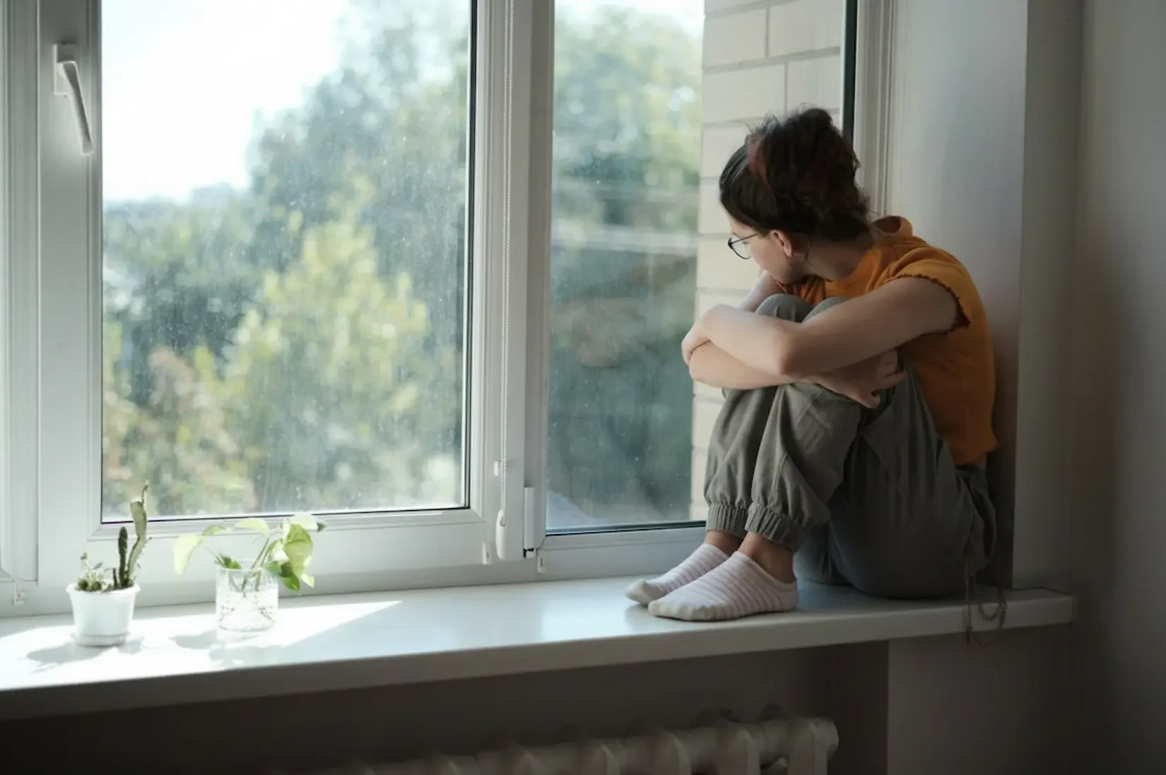 A young person sits reflectively on a windowsill, illustrating the quiet moments of an individual's mental health journey or the need for remote support.