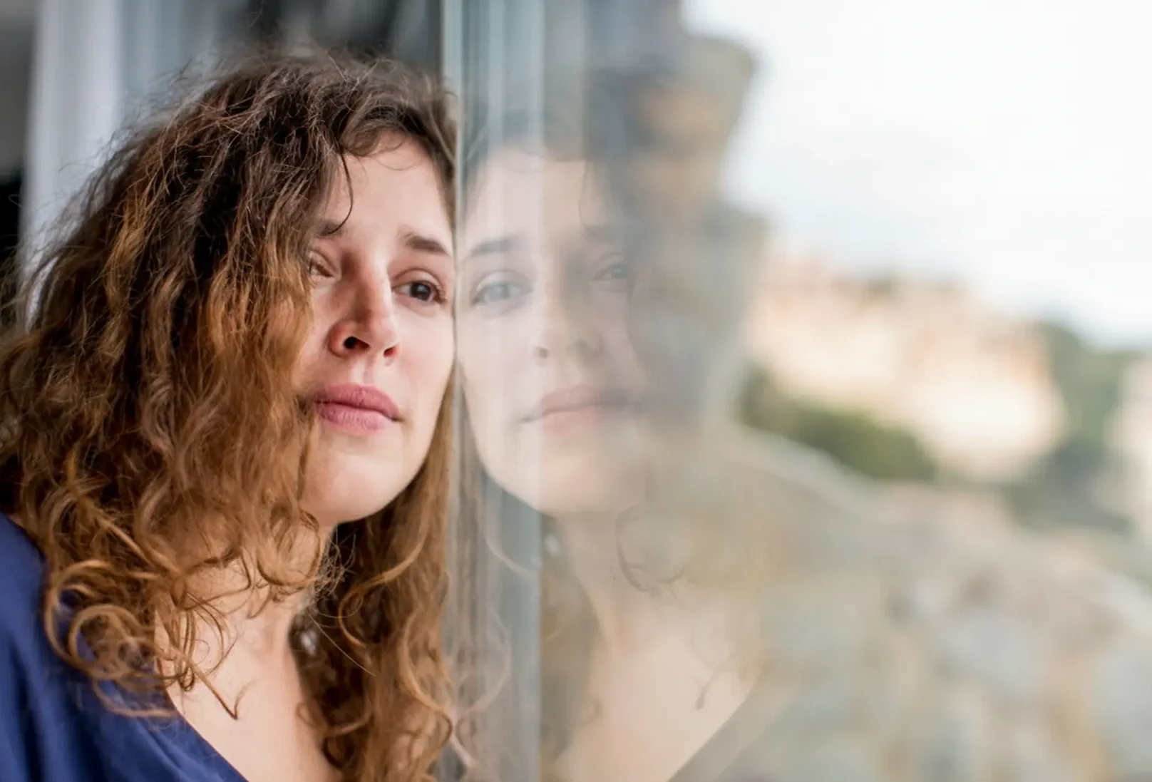 A woman with curly hair looking pensively out a window, reflecting the emotional journey and personal breakthroughs common in psychiatric treatment.
