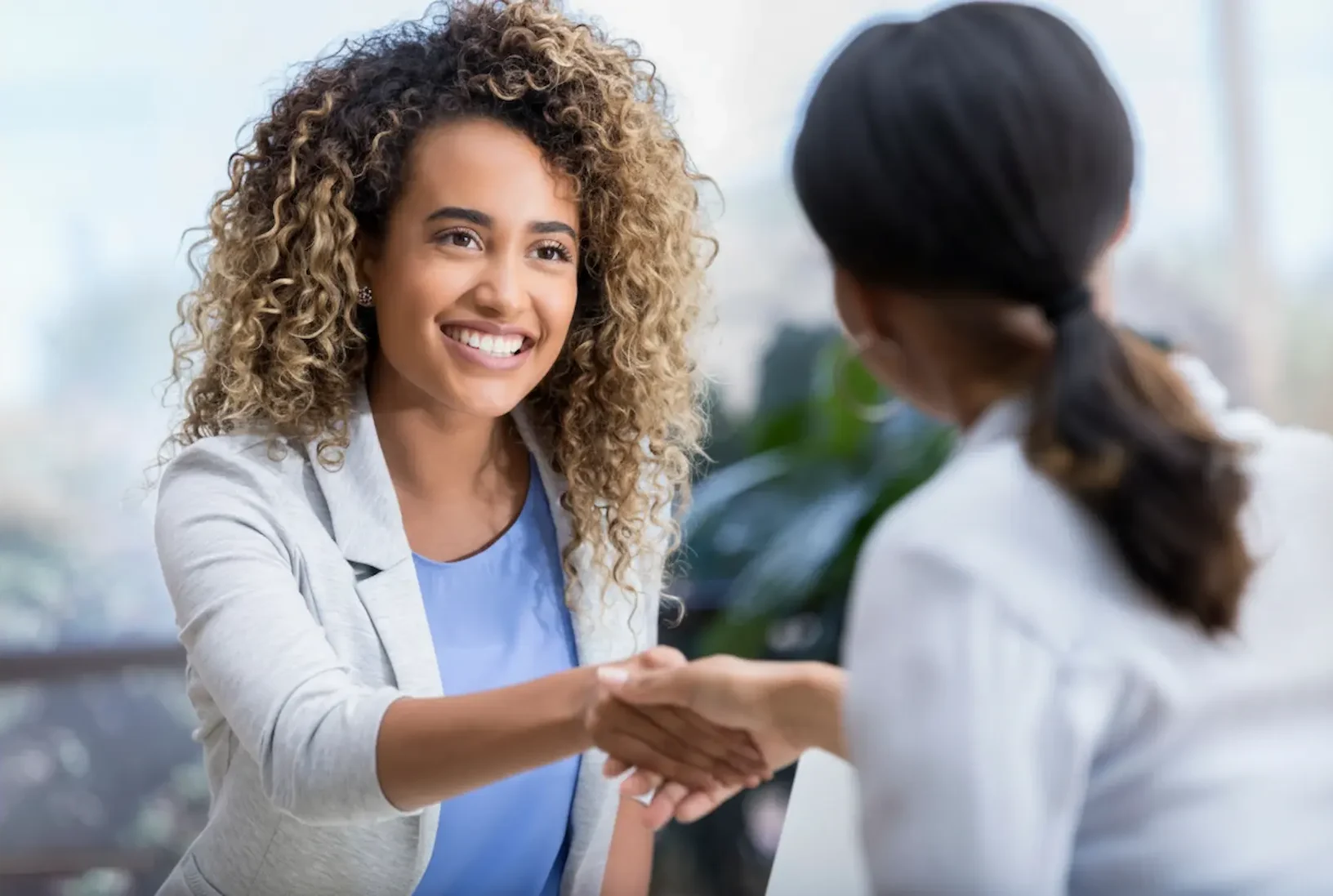 A smiling woman with curly hair shaking hands with a mental health professional, representing the start of a successful partnership with the right psychiatrist service.