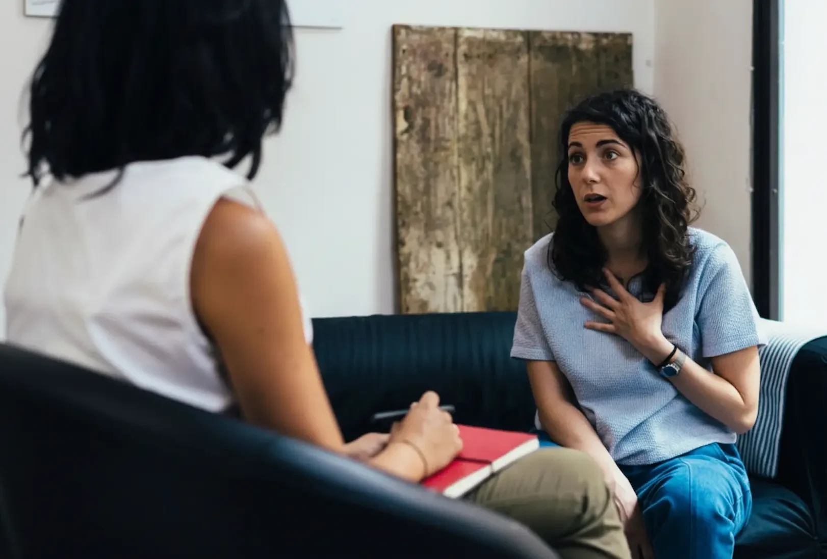 A woman expresses intense emotion during a psychiatric treatment session while a mental health professional takes notes in a red notebook.