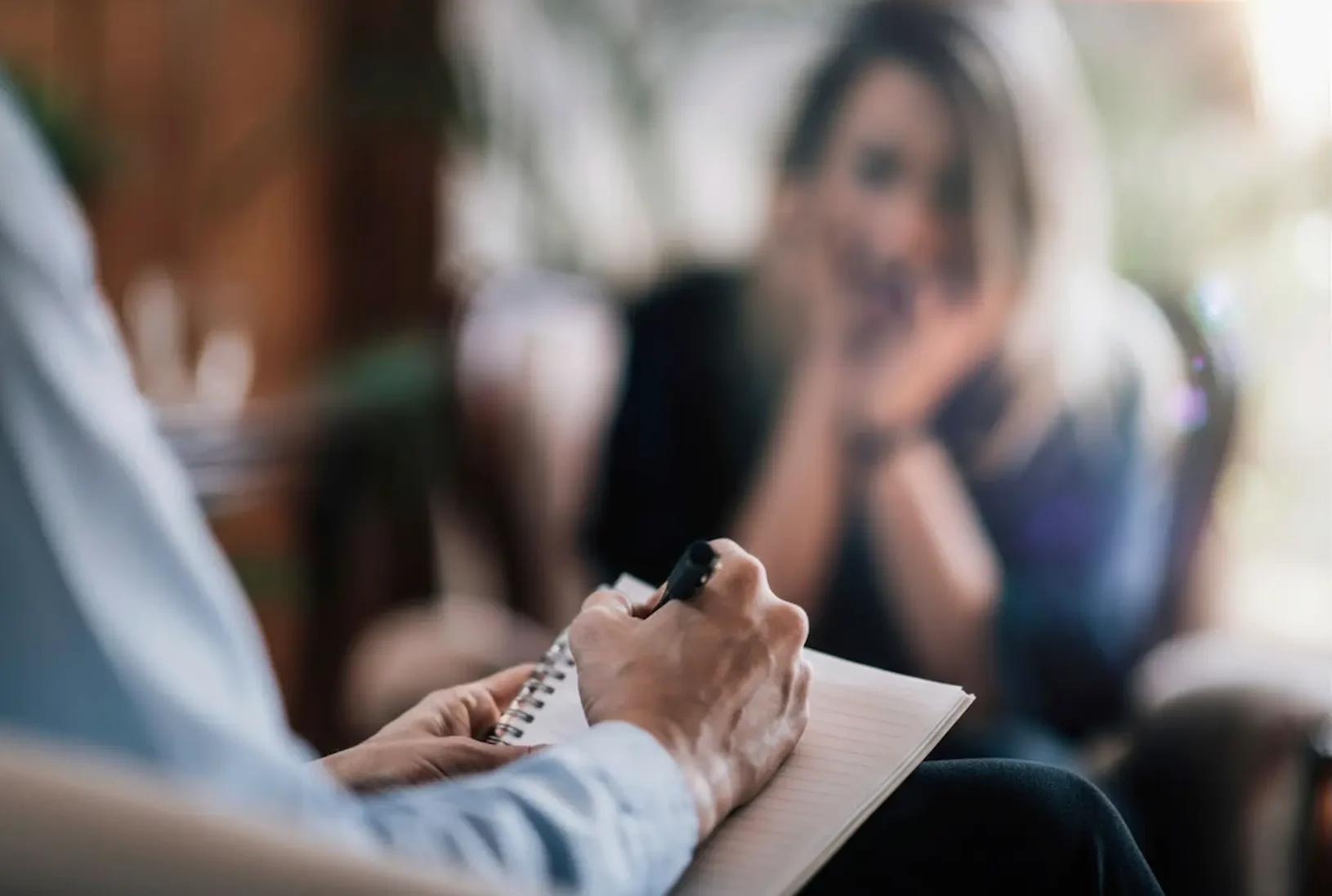 se-up of a healthcare provider taking notes during a psychiatric care session with a blurred patient in the background.