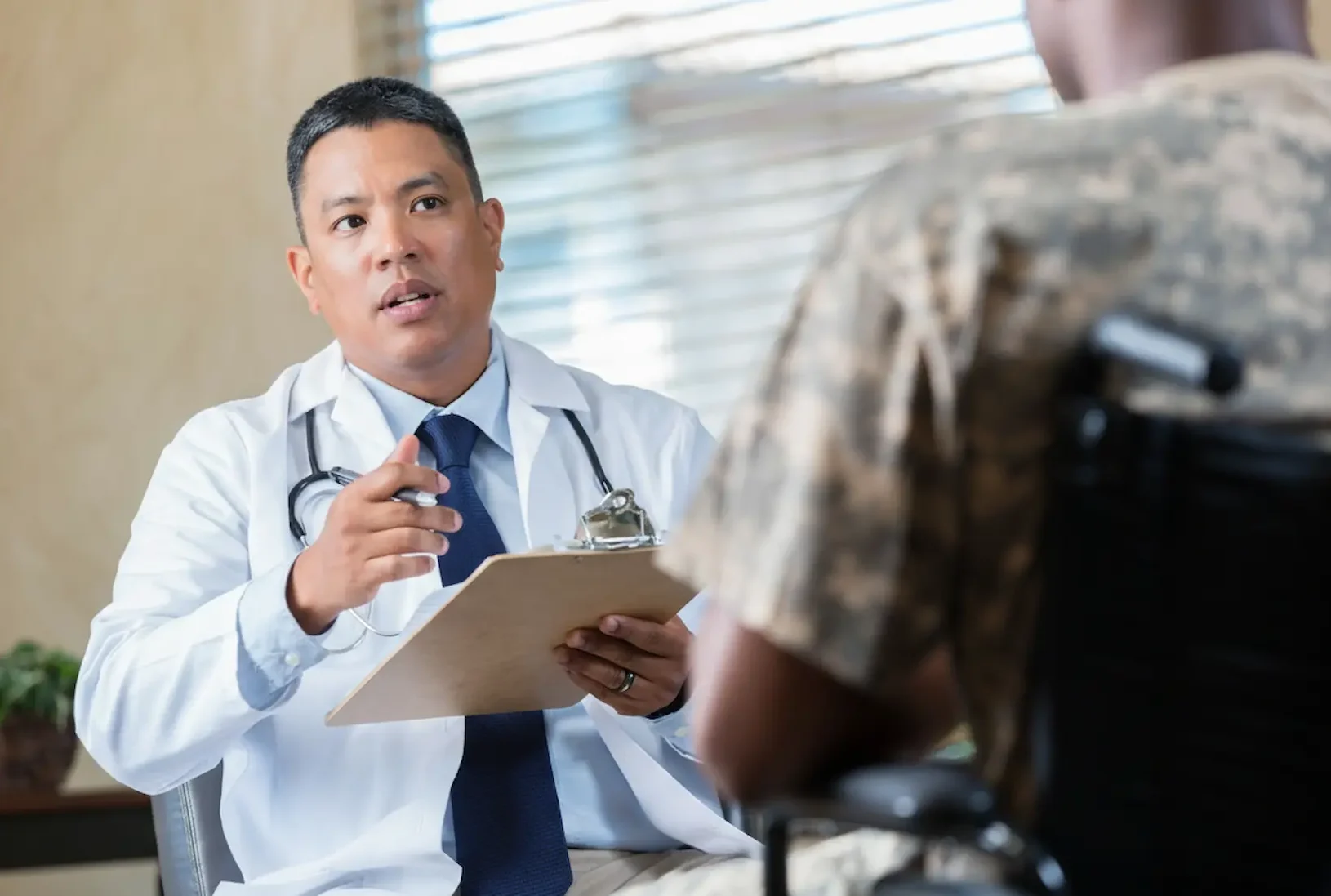 A male doctor in a lab coat conducting a psychiatric evaluation for a military veteran in a wheelchair.