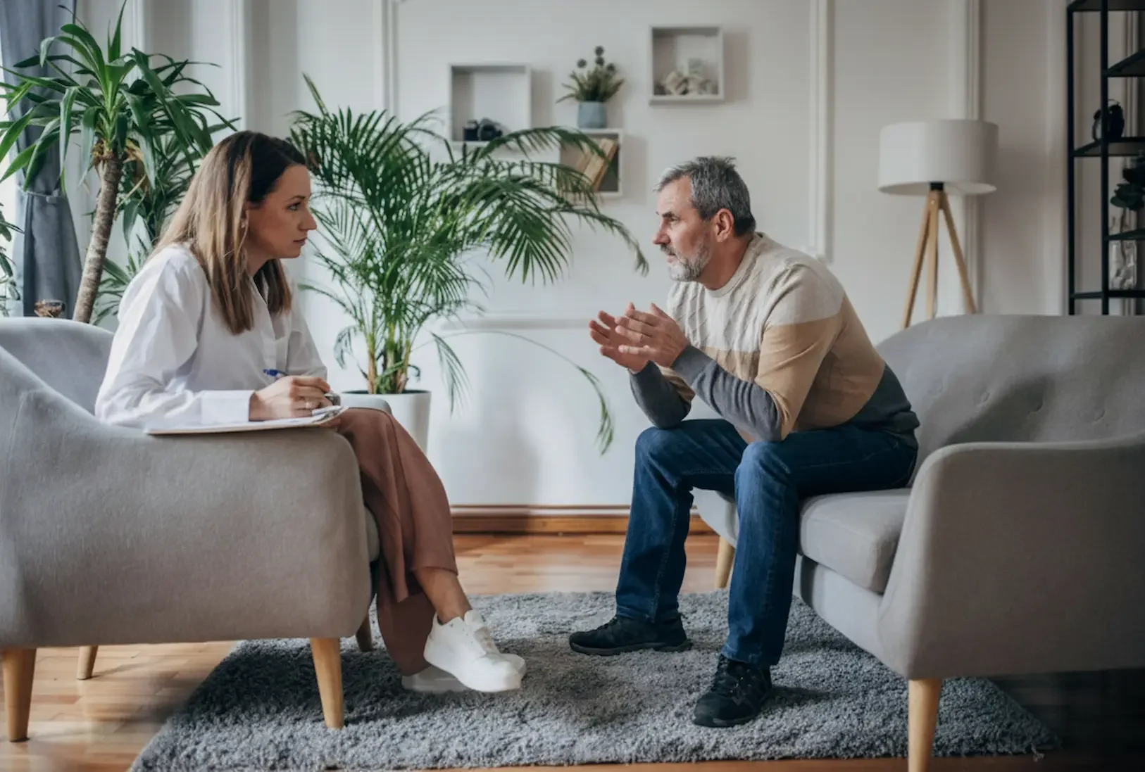 A female psychiatrist providing professional mental health services while listening to a middle-aged male patient in a bright, modern office setting.