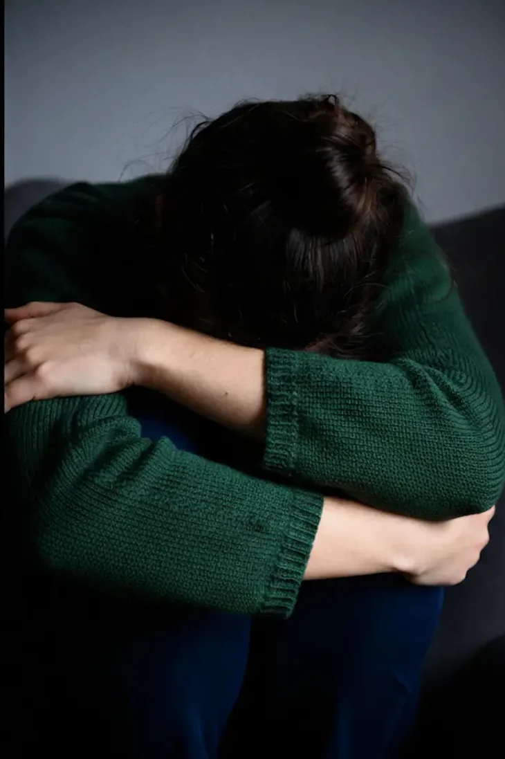 A person in a green sweater sitting alone with their head tucked into their arms, illustrating the emotional isolation associated with depression in adults.
