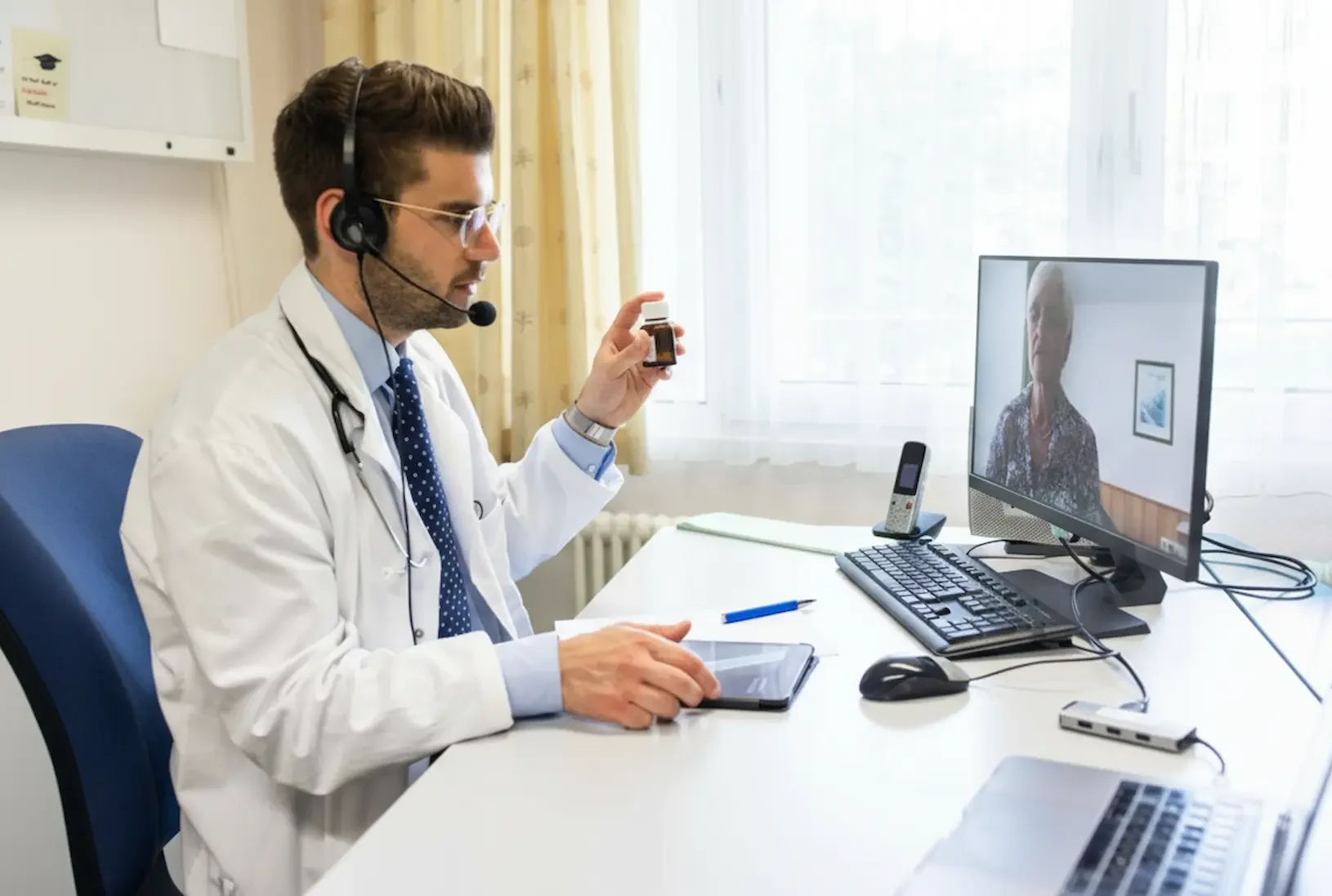 A male doctor wearing a headset holds up a prescription bottle to a computer screen during a virtual psychiatry session with an elderly female patient.