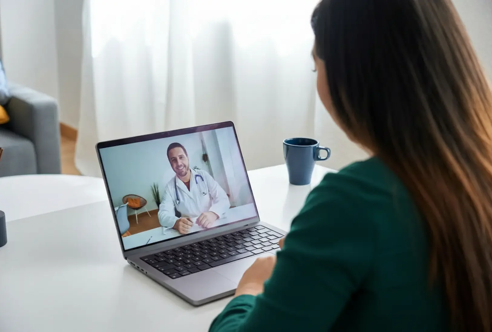 Close-up of a laptop screen showing a smiling doctor Providing telehealth Psychiatry care during a video call with a patient in a bright home office setting.