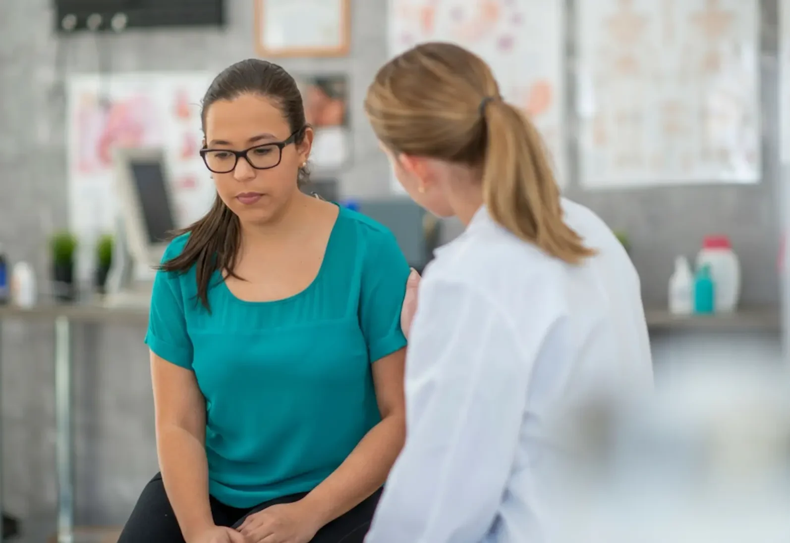 A young woman sits on an exam table during a psychiatric assessment, appearing pensive and downcast while a female clinician in a white lab coat offers supportive comfort with a hand on her shoulder in a clinical office setting.
