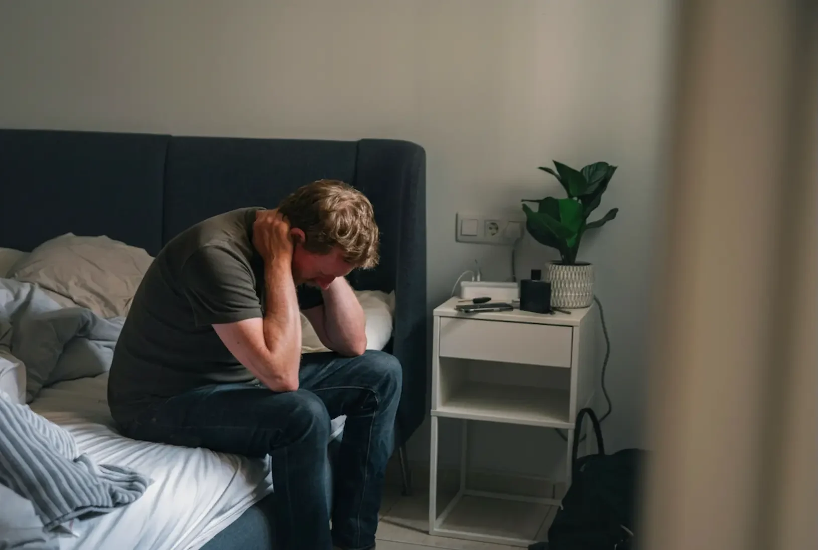 A man sitting on the edge of an unmade bed in a dimly lit room, holding the back of his neck and looking down while struggling with symptoms of mental illness.