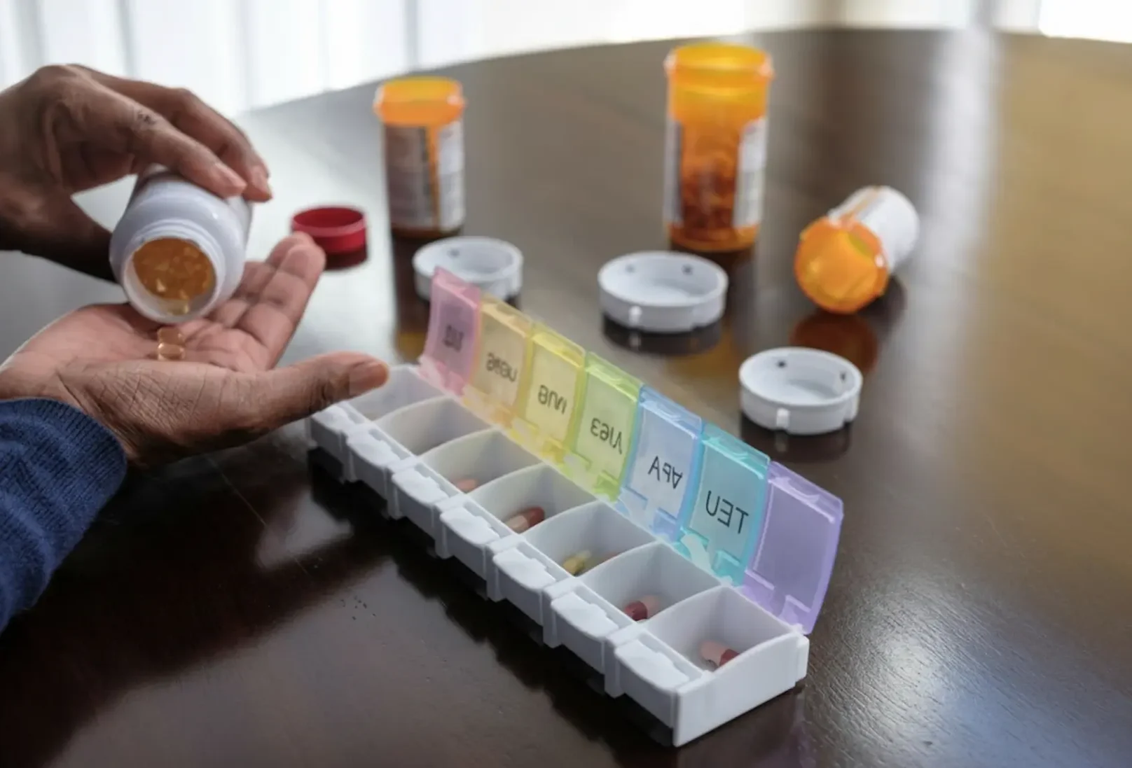 A person pouring capsules from a white bottle into their hand next to a colorful weekly pill organizer, a key tool for successful medication management.