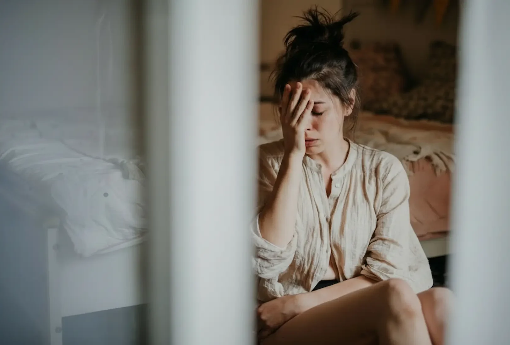 A woman sitting on her bedroom floor looking distressed and overwhelmed, holding her hand to her face while experiencing symptoms of an anxiety disorder.
