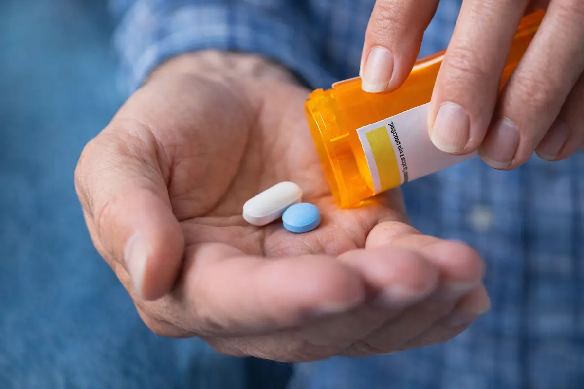 Close-up of a man’s hand holding two tablets with a prescription bottle, illustrating medication management and safe medication use.
