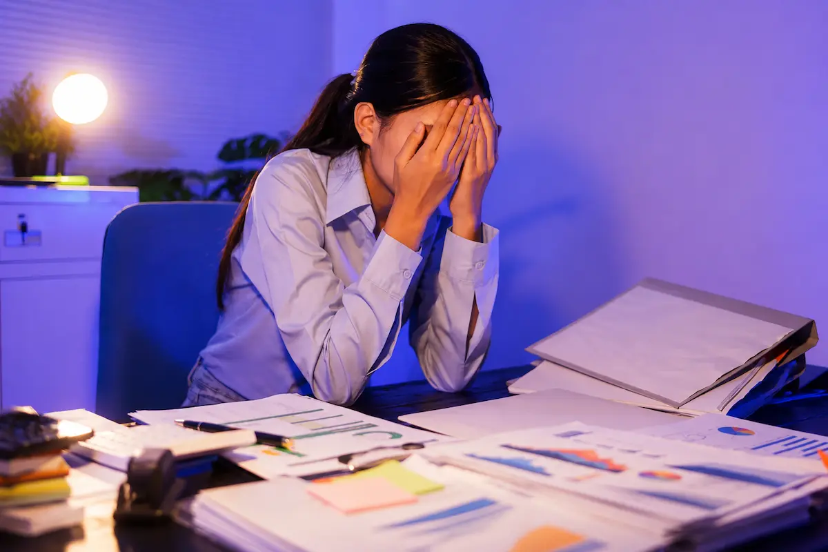 Woman experiencing adjustment disorder while sitting stressed at a cluttered office desk with paperwork and charts during late-night work.
