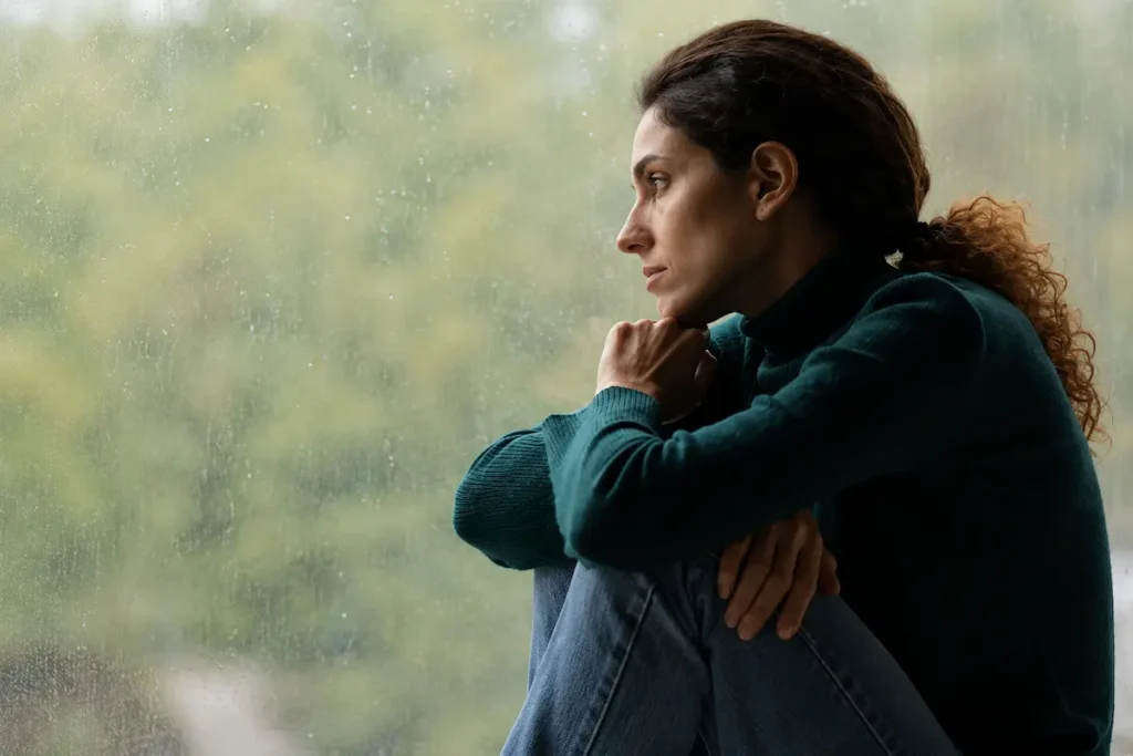 Woman sitting alone by a window in deep thought, representing the emotional challenges and symptoms associated with postpartum depression.